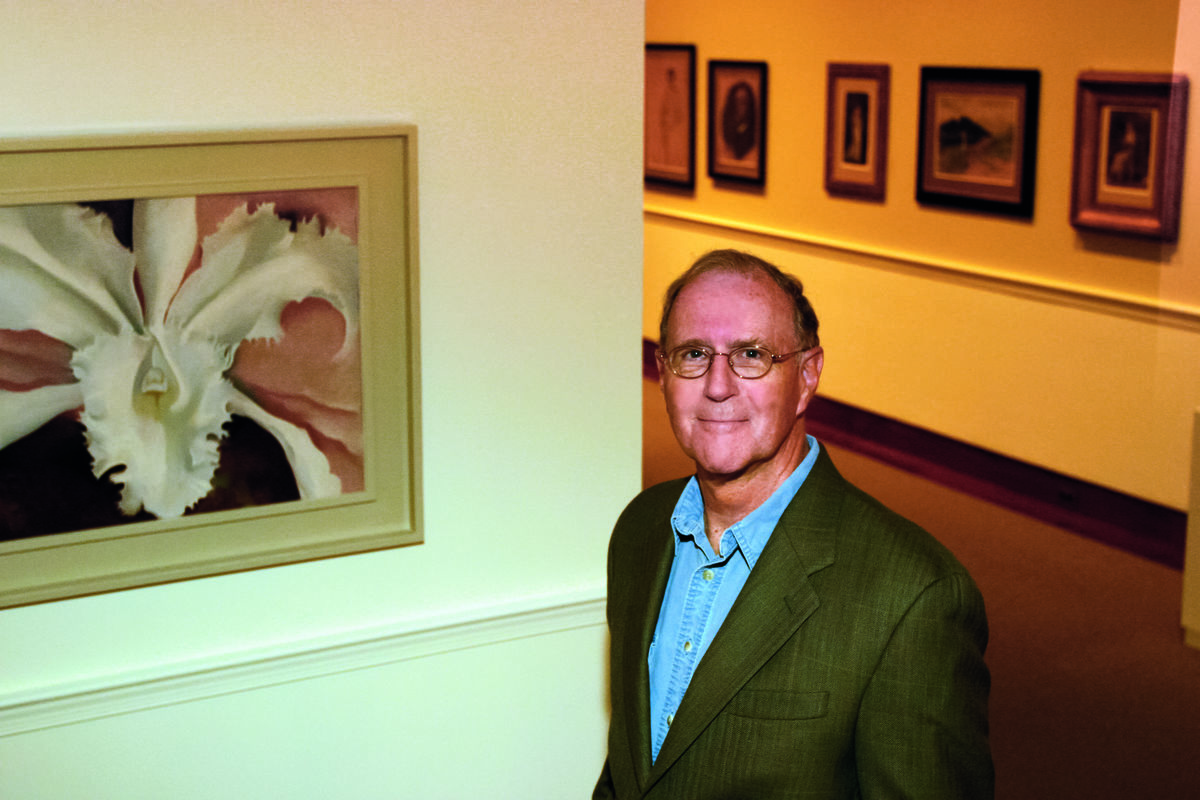 A light skinned man standing in a gallery next to a Georgia O'Keeffe orchid painting.